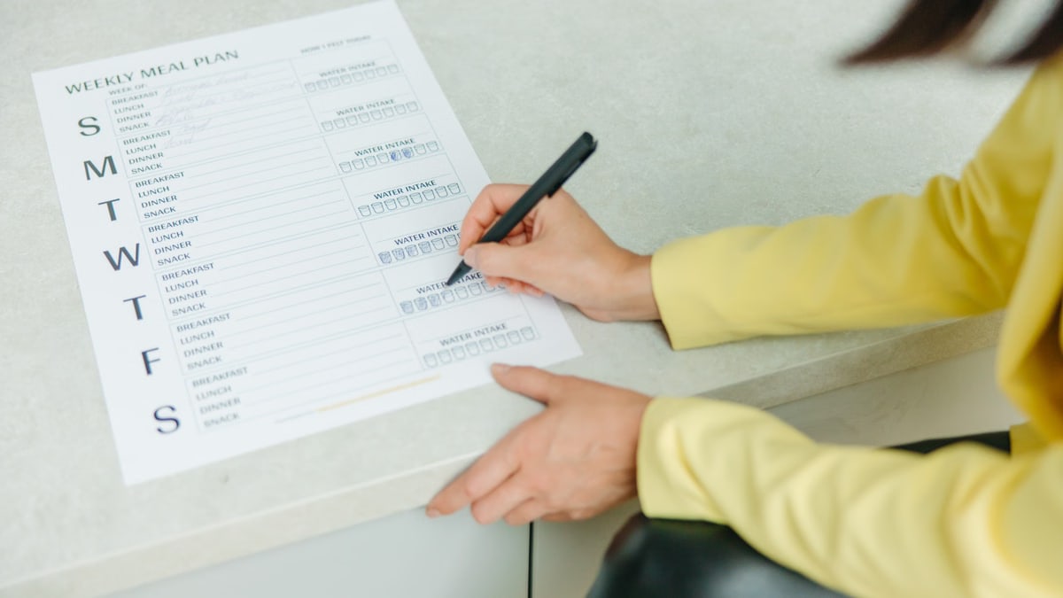 Woman writing on a weekly meal plan checklist indoors, focusing on diet organization.