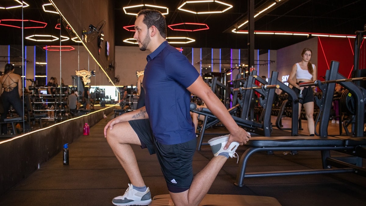 Mulher realizando exercício de agachamento em máquina de musculação com acompanhamento de personal trainer em academia moderna. A cena mostra treino de força com orientação profissional, foco na execução correta e segurança. Ambiente interno com equipamentos fitness, iluminação artificial e clima de disciplina e saúde.